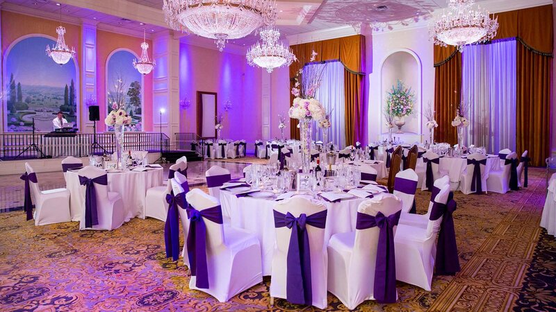 Ballroom. Six tables. White table clothes and seat covers. Purple napkins and chair accents with center pieces.